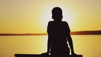 Silhouette of a boy sitting on bench at beautiful sunset over the river. Child is watching the sunset and simulates flying near the lake video