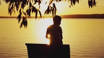 Small boy is sitting on a wooden dock at the river in the evening. Photo of a boy at sunset looking on the river and trying to find somebody or something in summer video