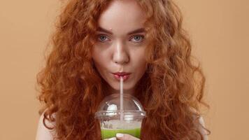 A woman with curly red hair sips from a smoothie cup, showing a vibrant green drink. The warm background highlights her bright smile and relaxed demeanor in a casual setting. video