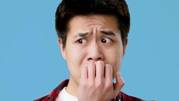 Asian young man exhibits a worried expression, biting his nails while glancing off to the side. The backdrop features a solid blue, adding to his anxious demeanor. video