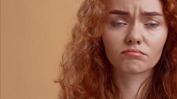 A young woman with curly red hair displays a look of sadness while standing in a studio. The warm background enhances her expression, capturing a moment of introspection. video