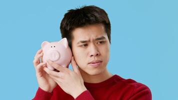 Asian young man in a red sweater curiously holds a pink piggy bank to his ear, listening intently as if expecting to hear something inside. The blue background adds a playful touch to the moment. video