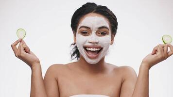 A joyful woman with a facial mask smiles while holding cucumber slices in each hand. Her bright expression embodies a fun and refreshing skincare routine at home, promoting self-care and wellness. video