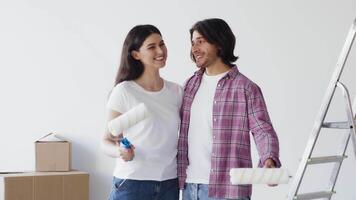 A couple stands in their new, empty home discussing plans to paint. The woman holds a paint roller while smiling as the man gestures, both excited about their DIY project. video