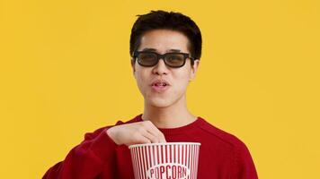 Asian young man wearing sunglasses and a red sweater is eating popcorn from a large striped container. The vibrant yellow backdrop adds a cheerful vibe to the activity. video