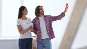 A couple stands in a well-lit, empty room, engaged in conversation about home renovation plans. The man gestures enthusiastically while the woman listens attentively, holding a notepad. video