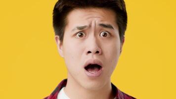 Asian young man displays an expression of shock and surprise, with his mouth open and eyes wide. Bright yellow backdrop enhances the intensity of his reaction, capturing a moment of strong emotion. video