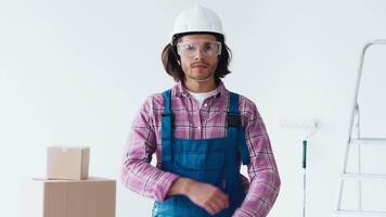 A construction worker wearing a hard hat and goggles stands with arms crossed in a bright workspace filled with cardboard boxes and tools, ready for the renovation project ahead. video