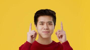 Asian young man wearing a red sweater stands in front of a bright yellow wall, pointing both fingers upward with a thoughtful expression, suggesting ideas or possibilities. video