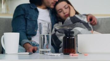 A couple is sitting on a couch at home, with the woman appearing concerned. They have medication and a glass of water on the table, highlighting a moment of support. video