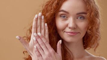 A woman with stunning curly red hair showcases her skincare routine at home, gently applying cream to her hand while smiling warmly at the viewer in a cozy setting. video