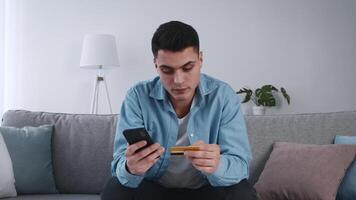 A young man is seated on a couch, focused on his smartphone while holding a credit card. The living room has a lamp and potted plant, creating a comfortable atmosphere. video
