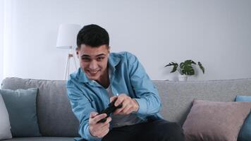 A young man sits comfortably on a couch, focused on gaming with a controller in hand. The modern living room features soft lighting and houseplants, creating a relaxing atmosphere. video