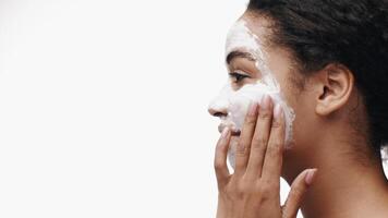 African American woman is gently applying a facial mask to her skin while smiling, showcasing her dedication to self-care and skincare. The bright, clean background emphasizes her glowing demeanor. video