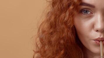 uma jovem mulher com vibrante encaracolado vermelho cabelo goza uma refrescante beber através uma canudo, com uma neutro bege fundo fornecendo uma caloroso ambiente este luzes dela brincalhão expressão. video