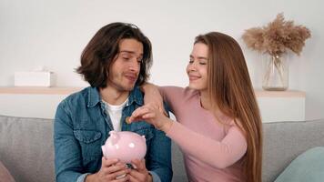 A young couple smiles at each other while holding a piggy bank in their cozy living room. They seem engaged in a light-hearted conversation about saving money together. video