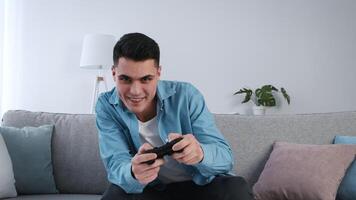 A young man enjoys playing games on a comfortable couch, smiling while focused on his game controller. The bright living room features soft furnishings and a plant nearby. video