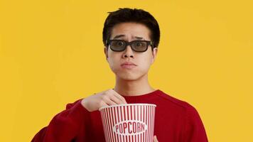 A young Asian man wearing glasses looks astonished while reaching into a large striped popcorn bucket, set against a vibrant yellow background. His expression conveys excitement. video