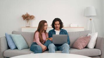 A young couple sits closely on a sofa, intently looking at a laptop screen while discussing their choices. One partner holds a credit card, indicating they are shopping online. video