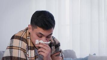 A young man, dressed in a blanket, appears to sneeze indoors, expressing discomfort. The soft light through the window creates a cozy, yet vulnerable atmosphere on a chilly day. video