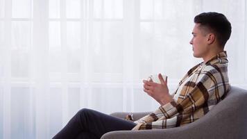A young man dressed in a plaid shirt sits comfortably on a couch, holding a remote control, enjoying a peaceful moment of relaxation in his cozy living room. video