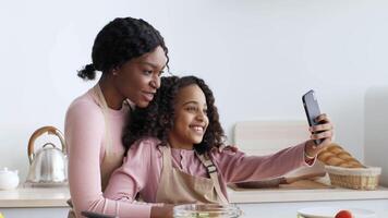 In a bright kitchen, black mother and her daughter are happily taking a selfie together. They enjoy cooking and sharing smiles, surrounded by ingredients and baked goods. video