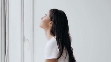 A young woman with long hair sits beside a large window, smiling softly while looking outside. Natural light fills the space, creating an uplifting atmosphere as she reflects. video