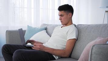 A young man is focused on his laptop while seated comfortably on a gray couch. Natural light pours in through large windows, illuminating the stylish living space. video