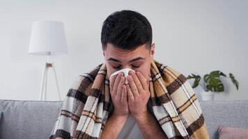 A man is seated on a couch, covering his face with a tissue. He appears unwell and is wrapped in a blanket, surrounded by a cozy indoor setting during wintertime. video