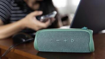 Close-up of a stylish green Bluetooth speaker placed on a table at home. The woman in the background sits at a table and works using a laptop and a smartphone. photo