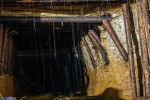 The dark and gloomy gold mine tunnel is supported by wooden beams. The walls of the tunnel are made of rough-hewn stone. The floor of the tunnel is covered in mud and water, gold mine photo