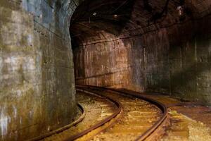 A dark, abandoned mine tunnel with rusty tracks, illuminated by a single light source, revealing its rough stone walls. It evokes a sense of history and mystery waiting to be discovered. photo