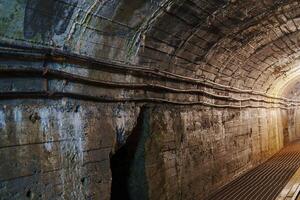 This dark tunnel features arched stone walls and dangling cables. Cracks in the ceiling provide minimal light, revealing the damp atmosphere and the sound of dripping water, cooper mine photo
