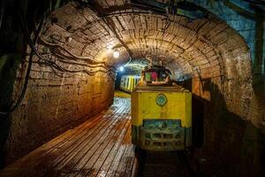 A rusty yellow mining train carries ore through a dimly lit tunnel. Dust particles float in the air as a single light illuminates the train, revealing the rugged stone and brick walls, cooper mine photo