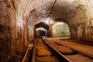 This image features an old dark mine tunnel with rusty railroad tracks. The rough stone walls create a spooky atmosphere, inviting exploration into the depths of the unknown. photo