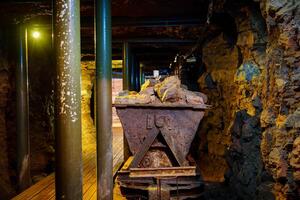 The metal cart, showing signs of rust, rests on aged wooden tracks in an abandoned mine shaft. The rough stone walls and dirt-covered floor enhance the atmosphere of neglect and history. photo