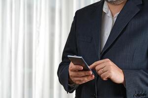 Confident businessman in a tailored suit actively using his smartphone for communication while standing in a spacious, modern office, showcasing professionalism and focus on work. photo