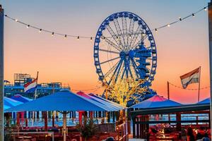 A stunning Ferris wheel stands tall at an amusement park, illuminated against a beautiful sunset sky, while a cozy restaurant with tables and umbrellas invites visitors to enjoy the view. Hague , Netherlands photo