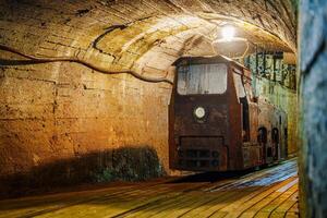 Old rusty train in a dark tunnel with a bright light at the end of the tunnel. The train is on wooden tracks in a tunnel carved out of solid rock. There is a wooden platform next to the tracks. photo