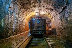 A rusty old mine locomotive sits idle in a dark mine tunnel with wooden tracks and stone walls. The locomotive is old, rusty, and covered in dust and is in poor condition and no longer in use, gold mine photo