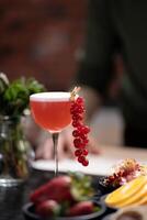Red cocktail garnished with fresh red currants in tall glass, placed on rustic table with various fruits and greenery in the background, evoking refreshing, vibrant, and summery atmosphere, copy space photo