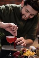 Bartender adding red currants to pink cocktail surrounded by fresh fruits and decorative elements on bar photo