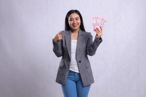 gesture beautiful young office female smile wide hand pointing and hold rupiah money wearing gray suit isolated on white background. for giveaway content, lifestyle, prize, event, competition photo