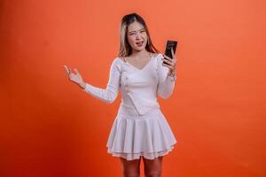 gesture young woman white top standing disappointed to camera hand holding mobile phone, cellphone and sideways isolated orange background. for communication, business, promotion, lifestyle concept photo
