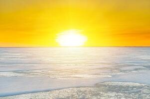 The setting sun casts a warm golden glow on the icy expanse of a frozen lake, while chunks of ice and snow create a textured foreground in this stunning winter landscape photography. photo