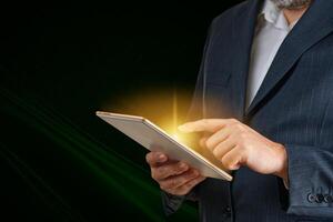 A confident businessman in a sharp suit interacts with a glowing digital tablet, showcasing the blend of technology and innovation in modern business against a dark background. photo