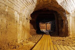 This image features a long underground tunnel with a vaulted ceiling made of stone blocks, dimly lit by a single light source at the end of the passageway. The walls are rough-cut stone. photo