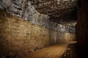 This image depicts a long underground stone tunnel featuring a wooden walkway on the right and rough stone walls illuminated by dim lights, showcasing medieval architecture. photo