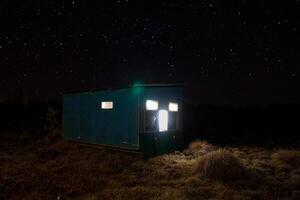 Wildlife Photo Hide in a grassy field, surrounded by tall trees. The warm glow from its windows contrasts with the cool night sky filled with twinkling stars, creating a serene atmosphere.