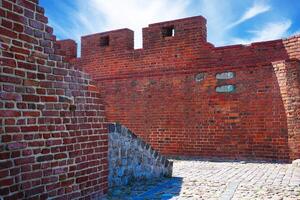 A historic red brick fortress wall with two turrets stands under a clear blue sky. A charming cobblestone street leads into the distance, featuring a hole in the larger turret. photo